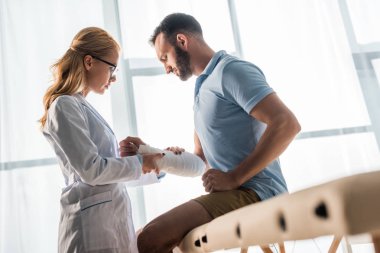 selective focus of beautiful orthopedist touching injured hand of bearded man 