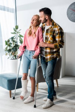 handsome bearded man looking at injured woman holding crutches at home