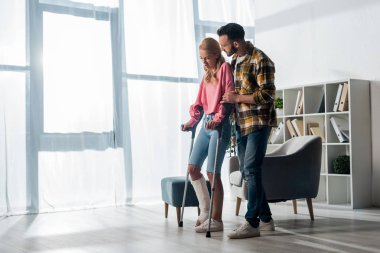 bearded man looking at injured woman holding crutches at home