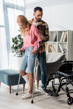 injured woman holding crutches while standing with man near wheelchair at home