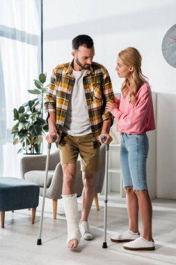 attractive woman looking at man holding crutches while standing at home 