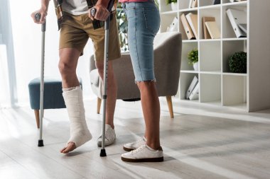 cropped view of woman standing near man holding crutches at home  clipart