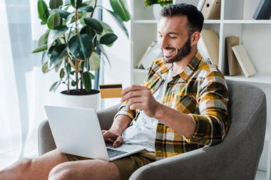 handsome and smiling man holding credit card while online shopping at home 