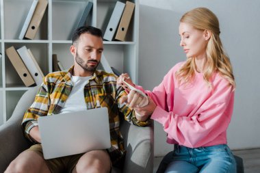 caring woman putting bandage on injured hand of bearded man sitting with laptop 