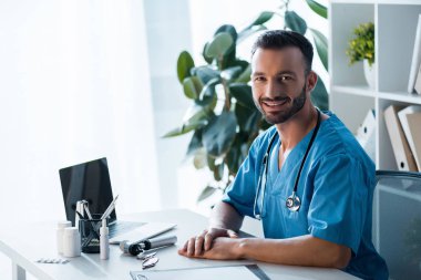 happy bearded doctor looking at camera near laptop in clinic 