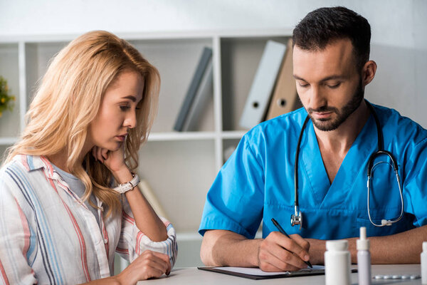 handsome bearded doctor writing prescription to woman in clinic 
