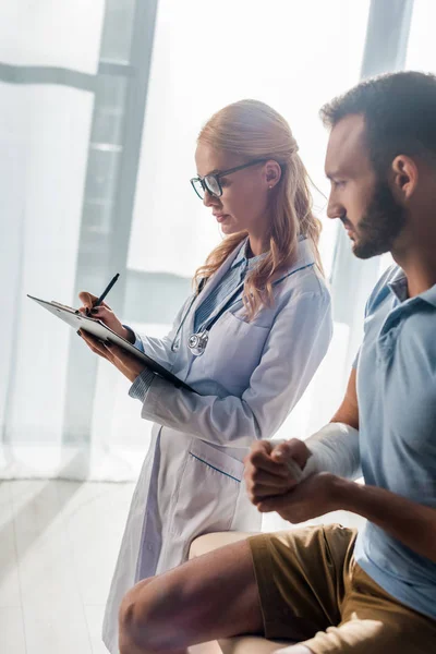 Attractive Orthopedist Writing Diagnosis Injured Bearded Man — Stock Photo, Image