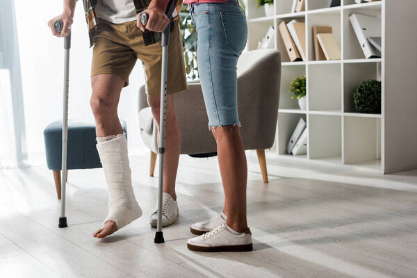 cropped view of woman standing near man holding crutches at home 