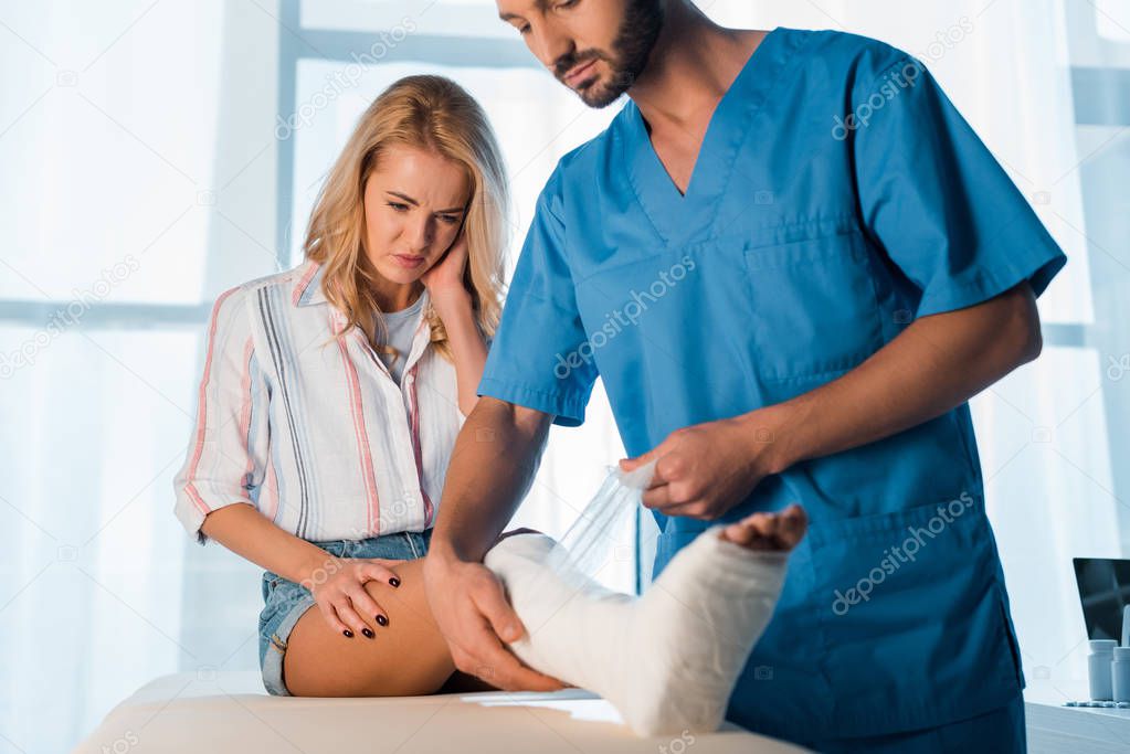 selective focus of bearded orthopedist putting bandage on fractured leg of woman 
