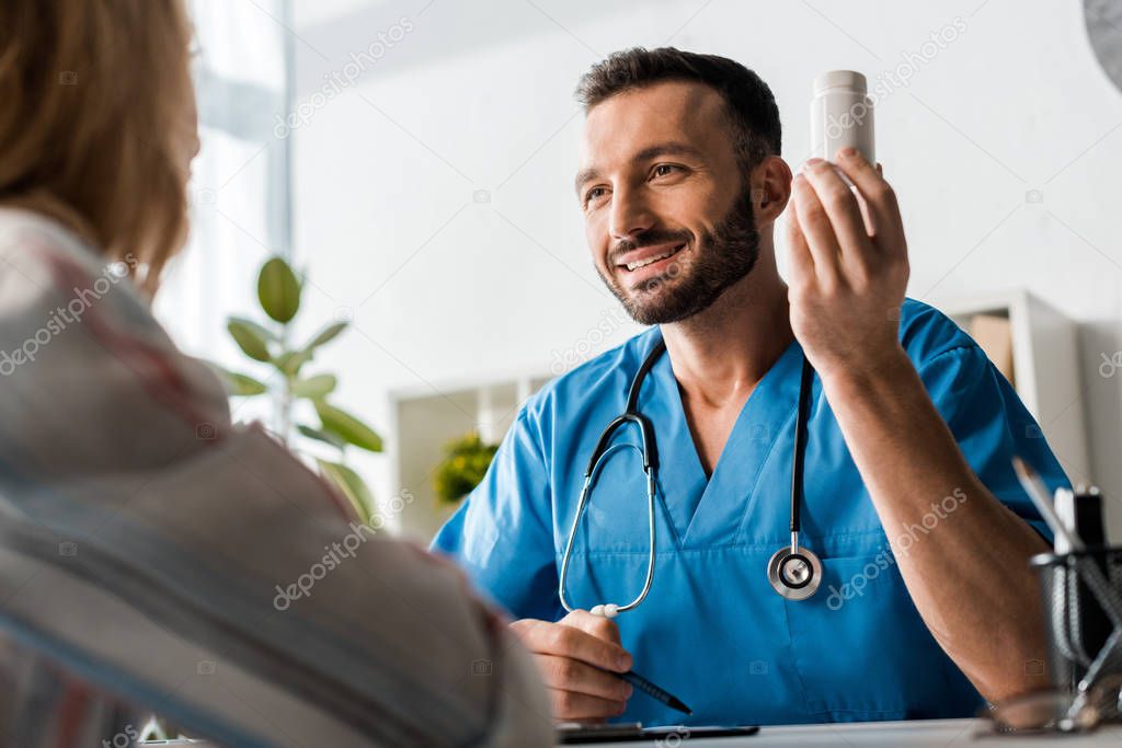selective focus of happy bearded doctor holding bottle with pills near woman