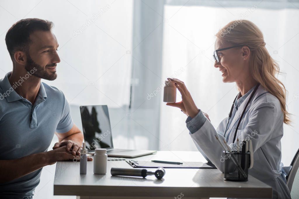 Cheerful doctor in glasses holding bottle with pills near bearded man