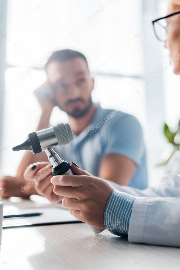 selective focus of doctor in glasses holding otoscope near handsome man in clinic 