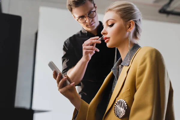 low angle view of Makeup Artist doing makeup to stylish model with smartphone on backstage