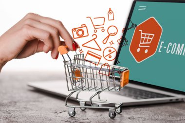 Dizüstü bilgisayarın yanında oyuncak alışveriş arabası tutan bir kadın ve beyaz, e-ticaret konseptini gösteren bir resim.
