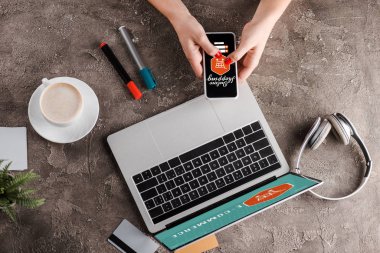 Bilgisayarın, kahve, bitki, kulaklık, kredi kartı ve elektronik ticaret konseptinin yanında online alışveriş mektupları olan akıllı telefon tutan kadının üst görüntüsü.