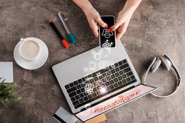 top view of woman holding smartphone near laptop, illustration, cup of coffee, plant, headphones and credit cards, e-commerce concept