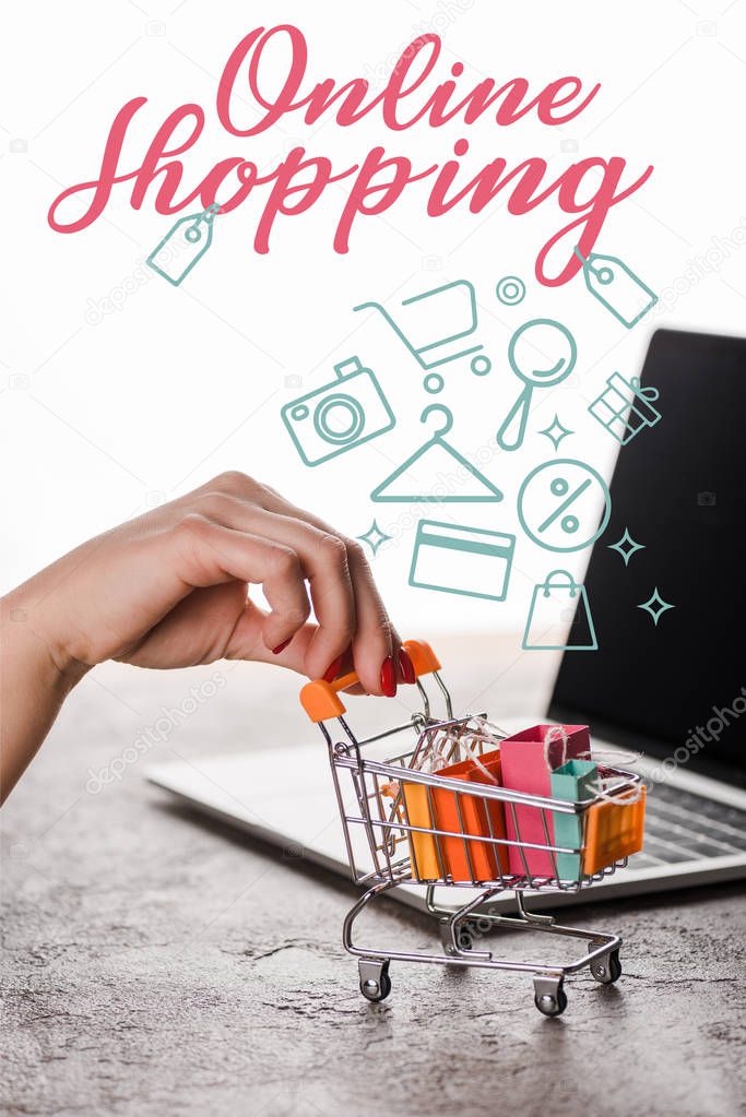 cropped view of woman holding toy shopping cart near laptop near online shopping letters on white, e-commerce concept