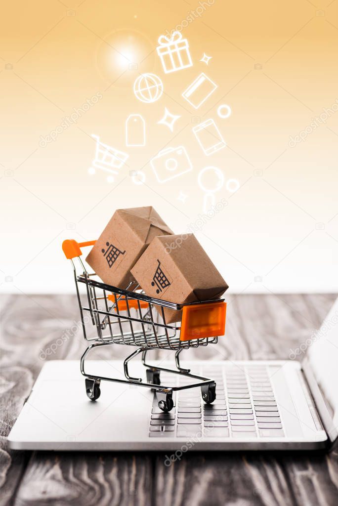 selective focus of toy shopping cart with small carton boxes on laptop keyboard near illustration on orange, e-commerce concept