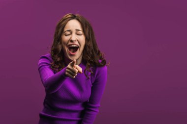 cheerful woman laughing with closed eyes and pointing with finger at camera on purple background clipart