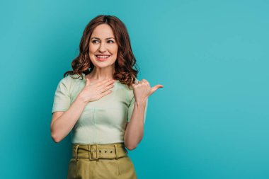 Şaşırmış, gülümseyen kadın başparmağıyla işaret ediyor ve mavi arka planda elini göğsüne koyuyor.