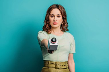 ciddi bir gazeteci mavi arka planda mikrofonu tutarken kameraya bakıyor