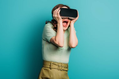 VR kulaklıklı korkmuş kız kafaya dokunuyor ve mavi arka planda çığlık atıyor.