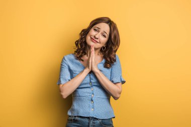smiling girl showing praying hands while looking at camera on yellow background clipart