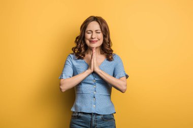 anxious girl showing praying hands with closed eyes on yellow background clipart