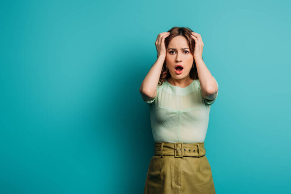 shocked woman touching head while standing with open mouth on blue background