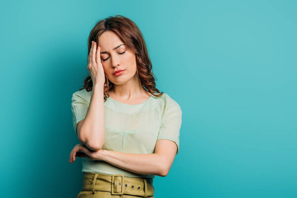 exhausted woman touching head while standing with closed eyes on blue background