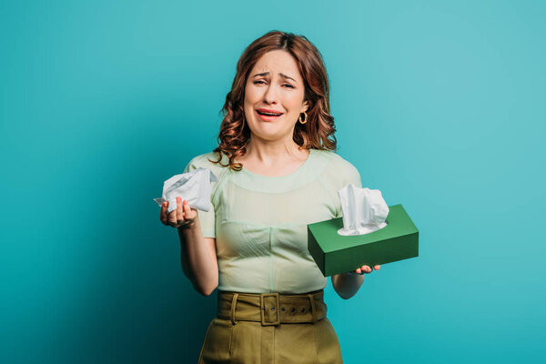 crying woman holding paper napkins and looking at camera on blue background
