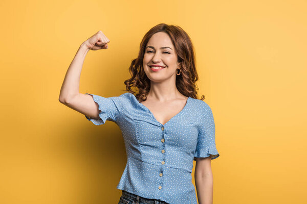 cheerful woman showing power while looking at camera on yellow background