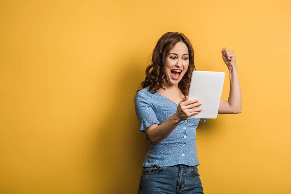 excited woman showing winner gesture while holding digital tablet on yellow background