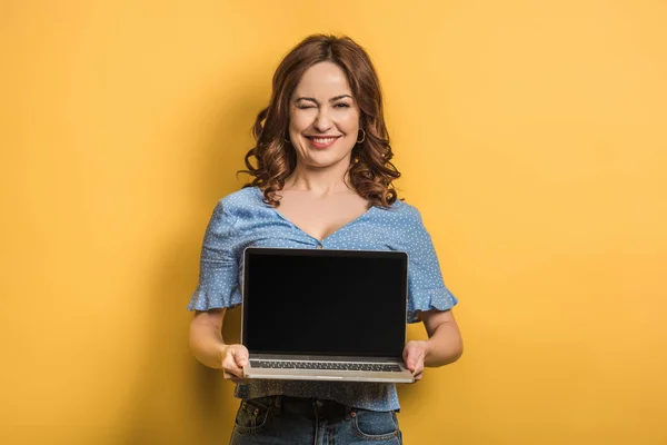 在黄色背景下拿着空白屏幕的笔记本电脑时 快乐的女人眨了眨眼 — 图库照片