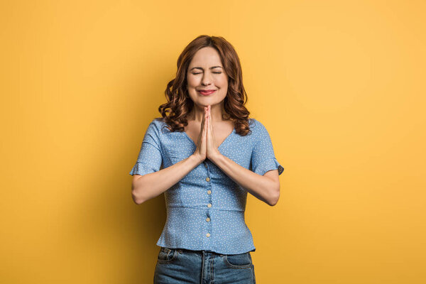 anxious girl showing praying hands with closed eyes on yellow background