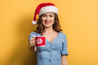 Noel Baba şapkalı mutlu kız sarı arka planda sıcak bir bardak içki tutuyor.