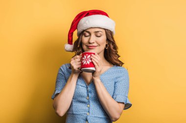 Noel Baba şapkalı neşeli kız elinde bir fincan sıcak içecek, sarı arka planda kapalı gözler.