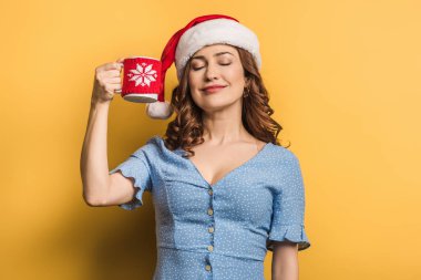 Noel Baba şapkalı mutlu bir kız elinde sıcak bir bardak, sarı arka planda gözleri kapalı.