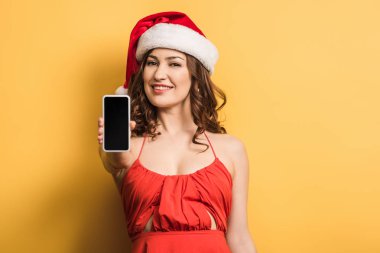 Noel Baba şapkalı gülümseyen kız sarı arka planda boş ekranlı akıllı telefonu gösteriyor.