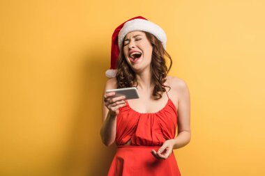 Sarı arka plandaki video çağrısında gülen Noel Baba şapkalı neşeli kız.