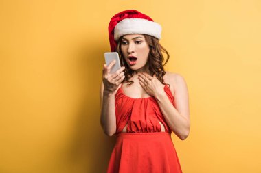 Şok olmuş Noel Baba şapkalı kız sarı arka planda akıllı telefona bakıyor.