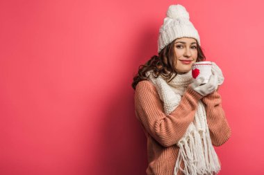 smiling girl in warm hat and scarf holding cup with heart symbol on pink background clipart