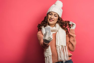 flirty girl in warm hat and scarf touching hair during video call on smartphone on pink background clipart