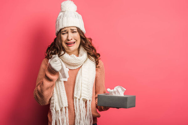 sad girl in warm hat and scarf crying while holding paper napkins on pink background