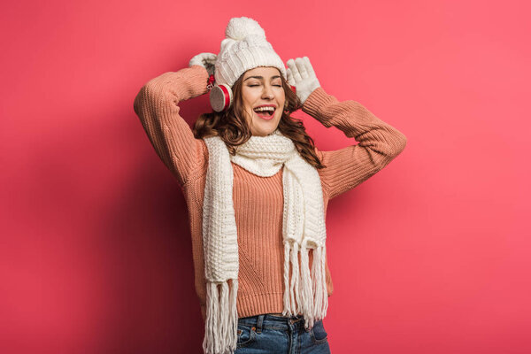 веселая девушка поет в беспроводных наушниках с закрытыми глазами на розовом фоне
