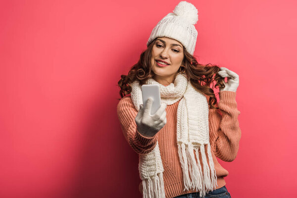 flirty girl in warm hat and scarf touching hair during video call on smartphone on pink background