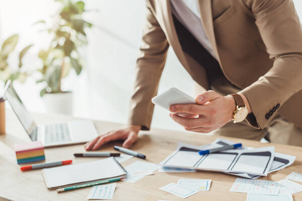Cropped view of ux designer using smartphone by website wireframe sketches on table
