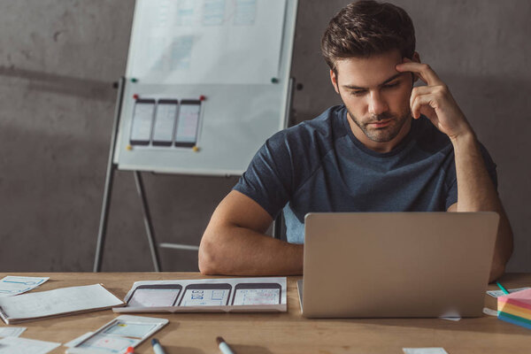 Selective focus of handsome ux designer using laptop beside wireframe sketches of mobile website in office