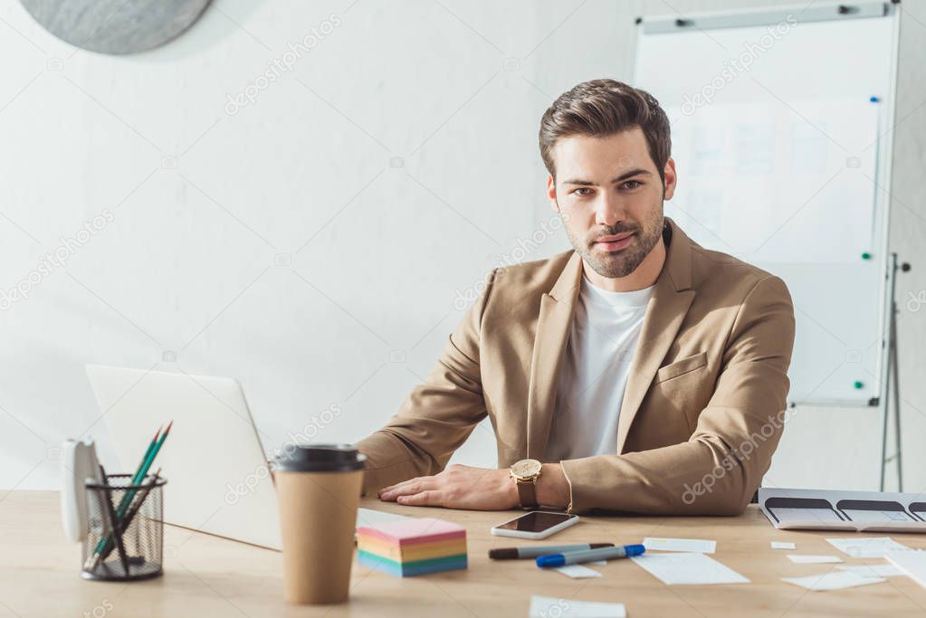 Handsome designer looking at camera while working with laptop and ux website templates in office