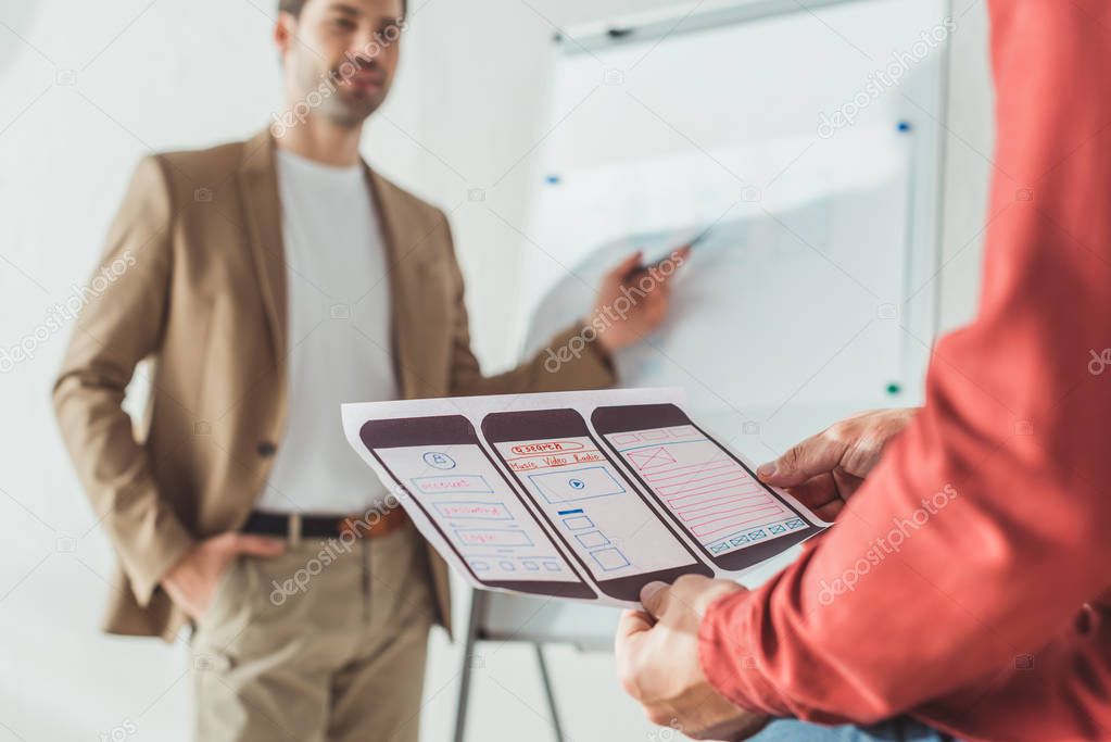 Selective focus of designer holding ux mobile application layout with colleagues beside whiteboard at background 